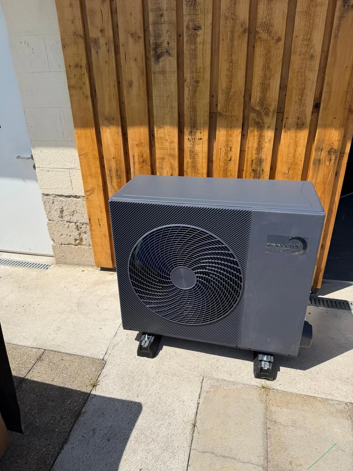 Unité extérieure de pompe à chaleur installée devant un bardage bois, sur terrasse pavée